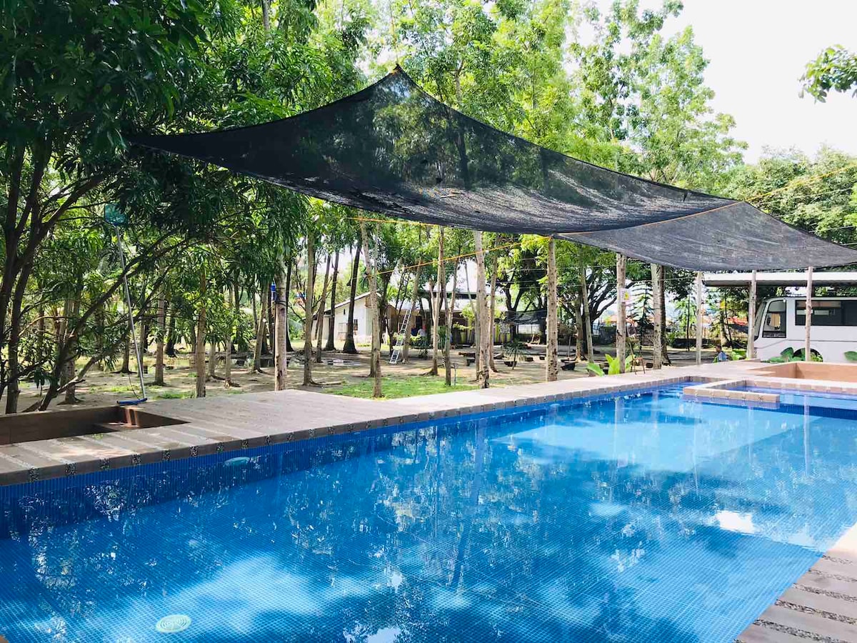 A private pool is surrounded by lush greenery, with a shaded area overhead providing respite from the sun. The pool's clear blue water reflects the trees, creating a serene and inviting outdoor space for relaxation.