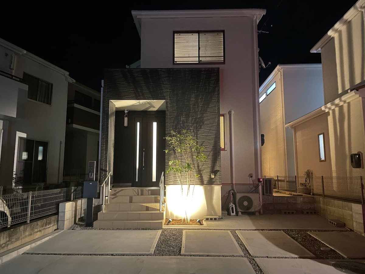 The exterior of a modern two-story house is illuminated at night, highlighting the sleek black front wall and entrance steps. A slender tree is positioned beside the steps, adding a touch of greenery. Nearby neighboring homes are also visible in the background.