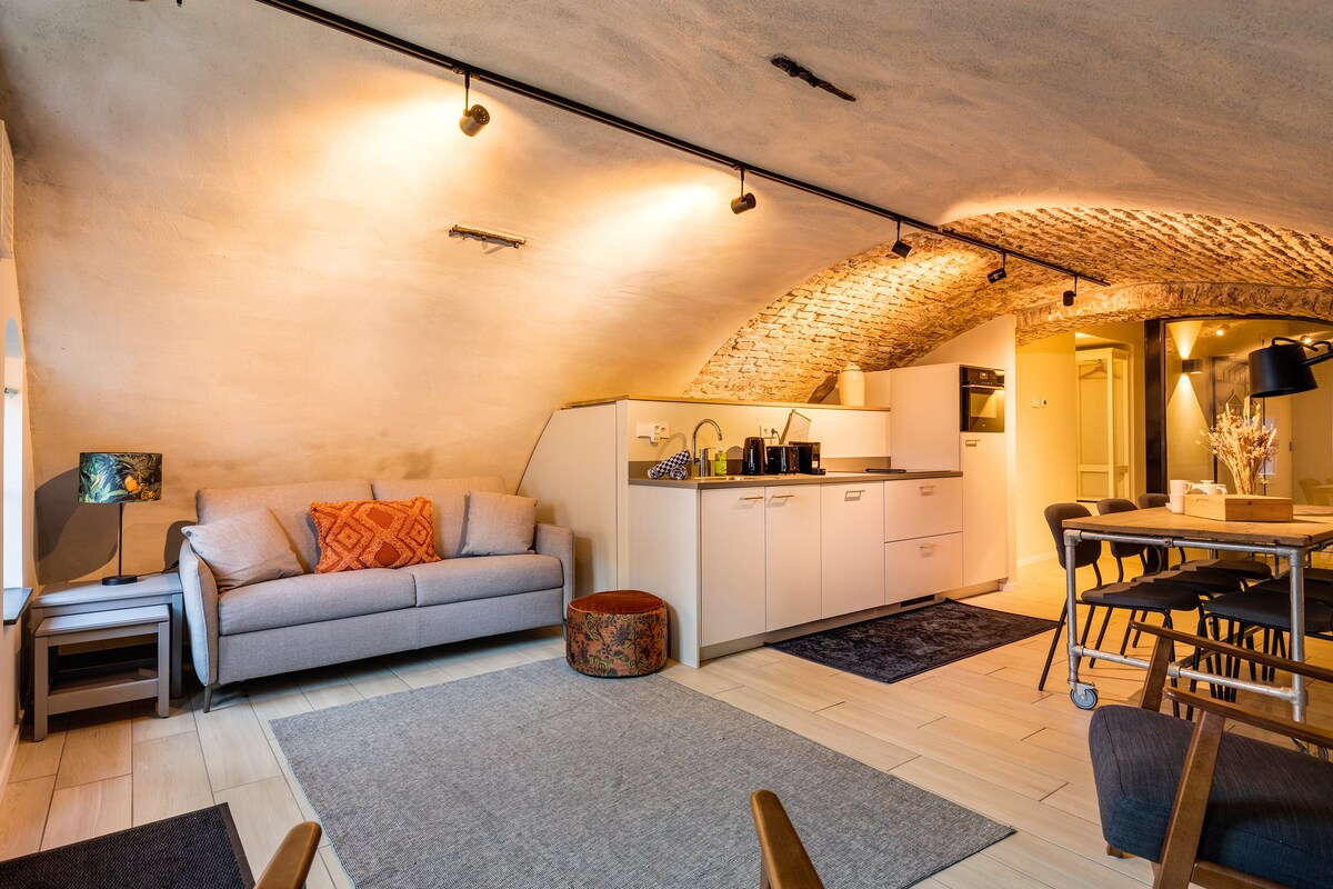 The open living area features a modern kitchen with sleek cabinetry and a cozy seating arrangement. A comfortable sofa is positioned near a decorative pouf. A dining table with chairs provides a welcoming space for meals, while softly lit arches create an inviting atmosphere.