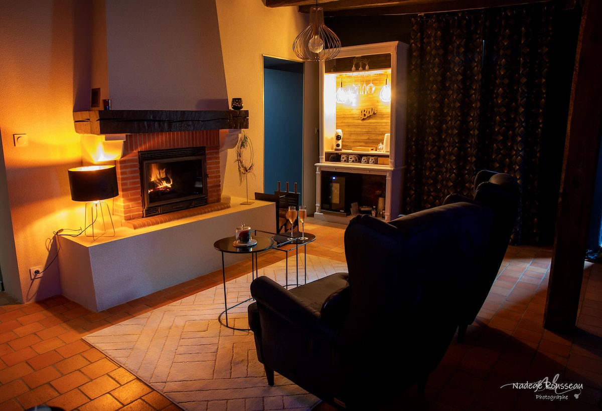 A cozy living area features a warm fireplace with a black mantel, complemented by a small round table. A plush armchair is positioned nearby, surrounded by a soft rug. A side lamp provides subtle lighting, enhancing the inviting atmosphere of the space.