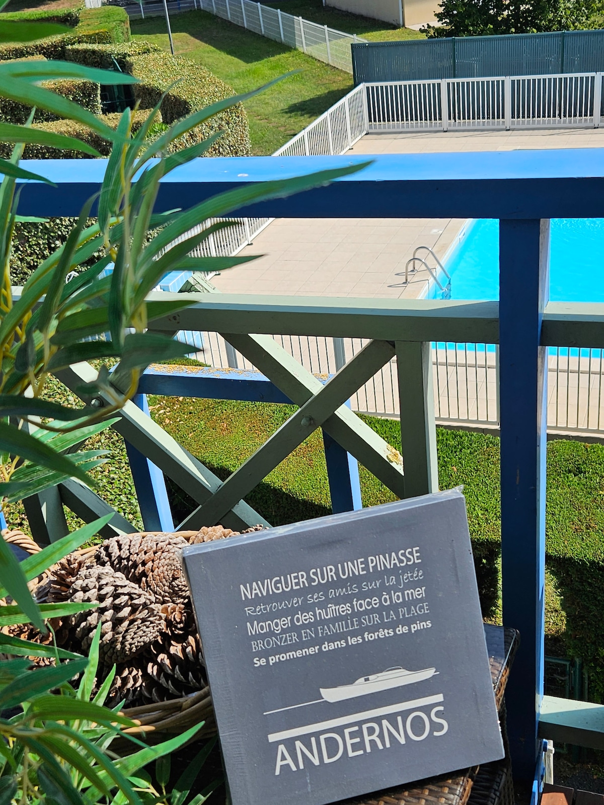 A wooden deck features a view of a swimming pool surrounded by landscaped greenery. A decorative sign offers details about local activities. Potted plants add a touch of nature, enhancing the inviting atmosphere of the outdoor space.