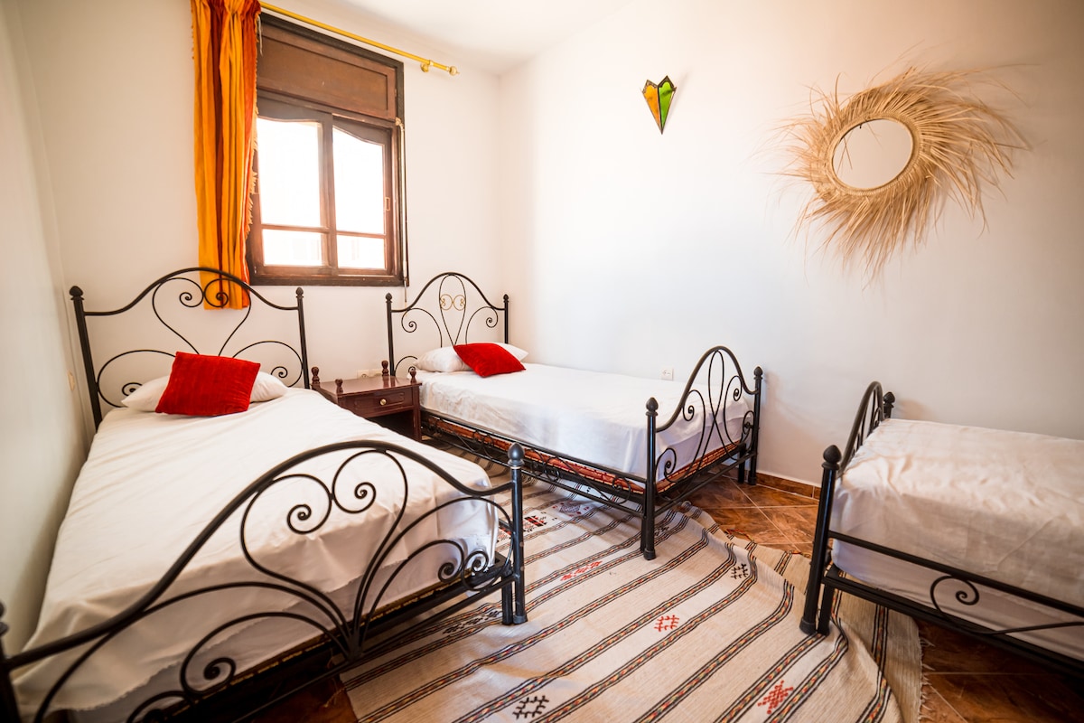 A bright room is furnished with three metal bed frames, two with matching red accent pillows. Natural light filters through a window adorned with curtains, casting soft shadows on a woven rug that covers the tiled floor.