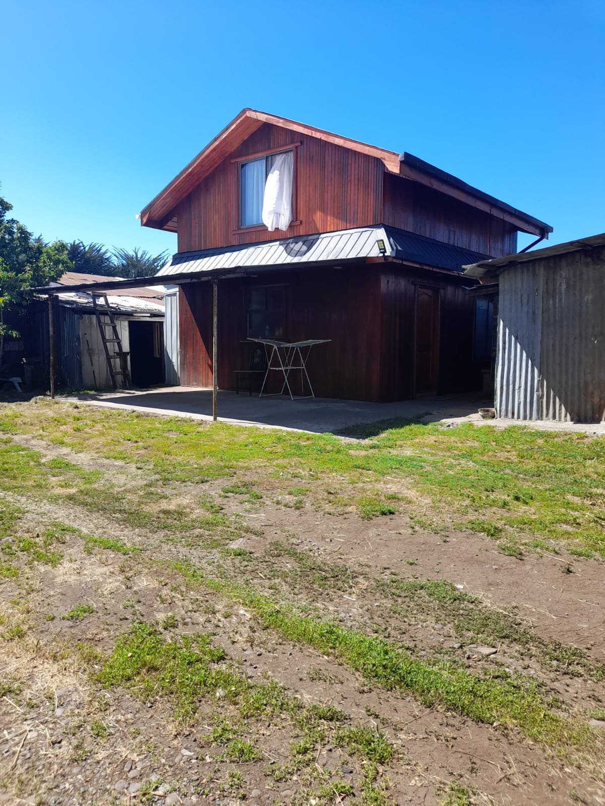 A two-story, wooden cabin is presented in this image, featuring a steeply pitched roof and a small front porch. A table and chairs are visible on the porch. Sunlight illuminates the surrounding grassy area, and a nearby shed can be seen on the left.