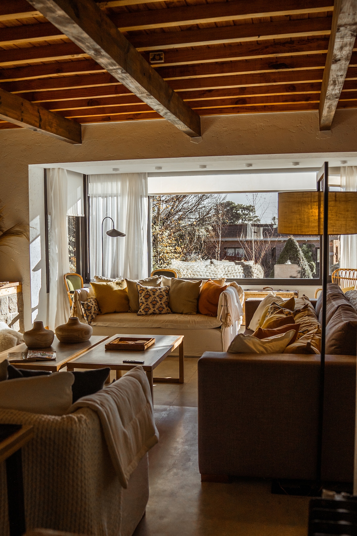The living area offers a spacious sofa adorned with decorative pillows, complemented by natural light filtering through large windows. Wooden beams highlight the ceiling, while rustic decor enhances the cozy ambiance. A mix of textures, including a soft throw and wooden furnishings, create a welcoming space.