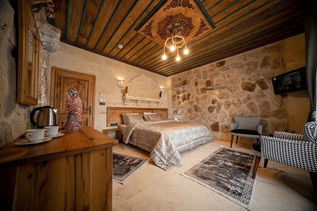 A spacious room features stone walls and natural wooden accents, creating a warm environment. A large bed with a patterned coverlet is centered, while a seating area with a patterned chair invites relaxation. Decor includes a decorative ceiling and a flat-screen TV.