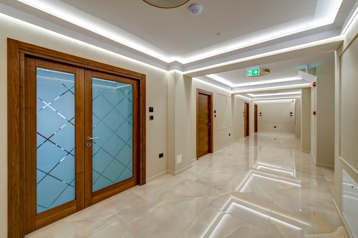 A well-lit hallway is lined with wooden doors featuring glass panel accents. The smooth, glossy floors reflect the ambient light from the recessed ceiling fixtures. The walls display a neutral color palette, enhancing the spacious and modern feel of the corridor.