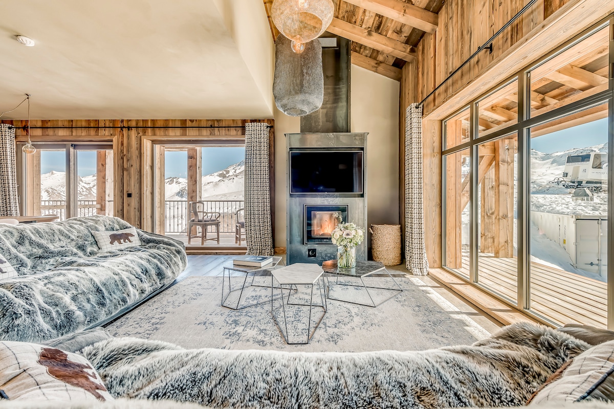 A cozy living area features plush, fur-covered seating arranged around a modern coffee table. A fireplace is positioned beneath a metallic accent, adding warmth. Large glass doors provide stunning views of the snowy mountains and open to a wooden balcony, inviting natural light into the space.