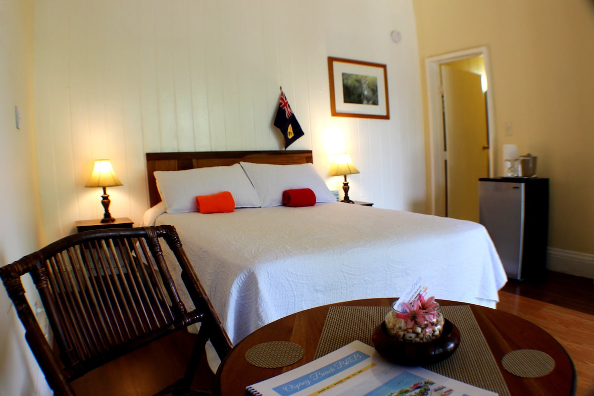 A cozy private room features a queen-sized bed dressed in white linens, complemented by two brightly colored pillows. A small round table with a decorative centerpiece sits next to a wooden chair. Soft lighting is provided by bedside lamps, enhancing the inviting ambiance.