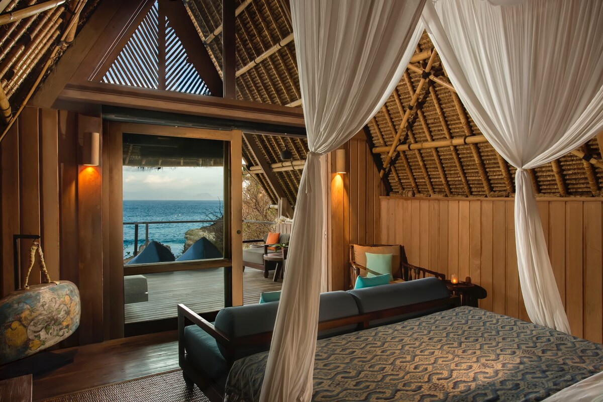 An inviting sleeping area features a spacious bed draped in elegant mosquito netting. Large glass doors open to a private terrace, showcasing ocean views. The room is constructed with natural materials, revealing bamboo accents and warm wooden walls, enhancing the tranquil atmosphere.