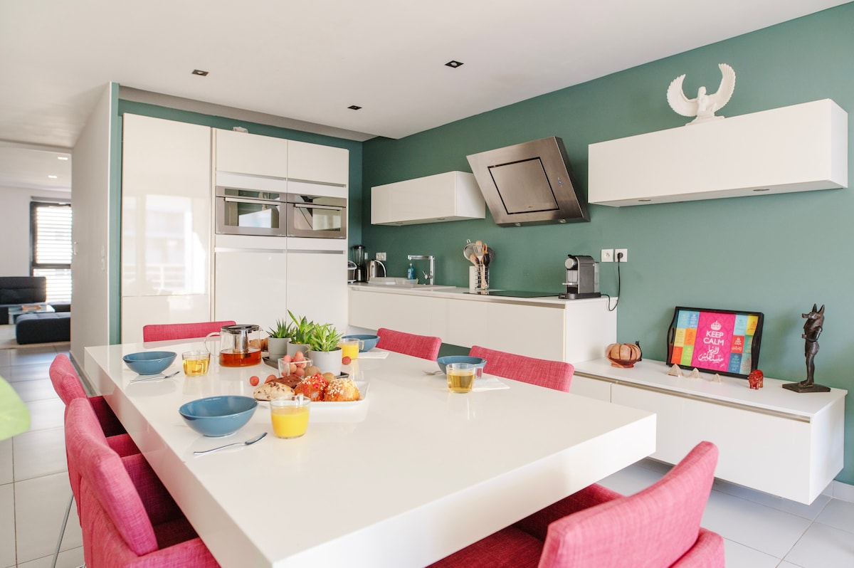 A modern kitchen is depicted, featuring sleek white cabinetry and a dark green accent wall. A large white table is surrounded by pink chairs, set with dishes and meal preparations. Appliances are neatly integrated, contributing to a contemporary and functional space.