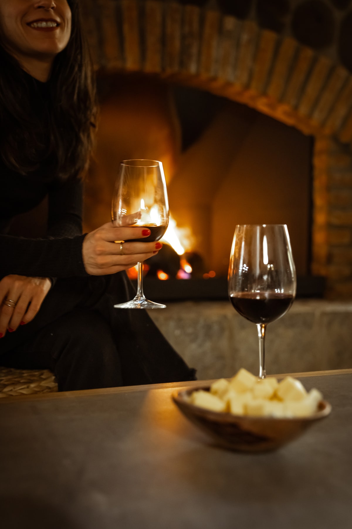 A cozy living space is depicted, featuring a warm fireplace in the background. Two glasses of red wine are placed on a table, alongside a bowl of cheese. Soft lighting enhances the inviting atmosphere of the room.