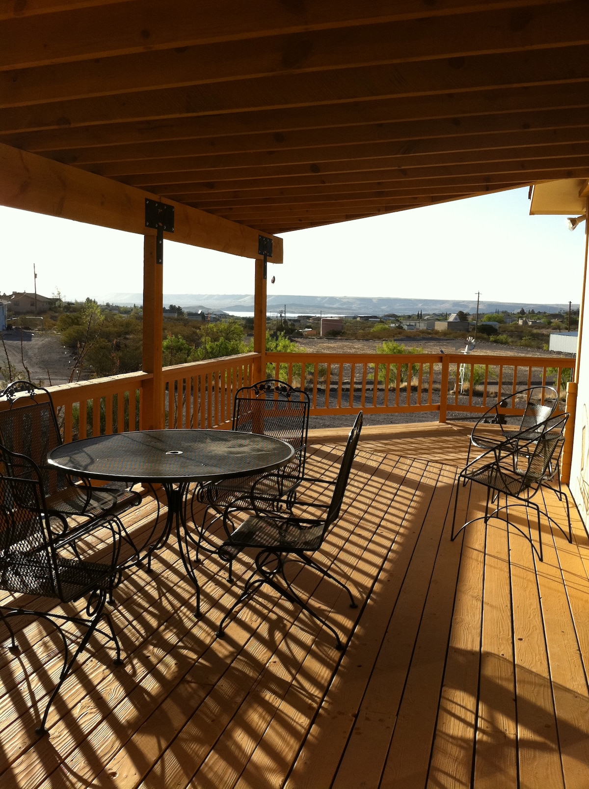 A spacious wooden deck is featured, furnished with a circular table and several black metal chairs. Sunlight casts shadows across the deck, highlighting the open view of the surrounding landscape and distant horizon.