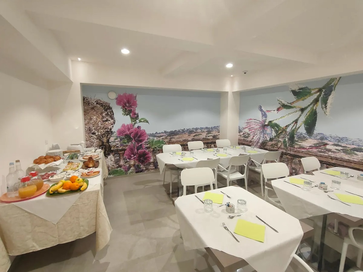 A bright dining area is presented with white tables and chairs, arranged for guests. A buffet selection of fresh fruits and pastries is visible, complemented by a large floral mural depicting vibrant natural scenery on the wall, enhancing the welcoming ambiance.