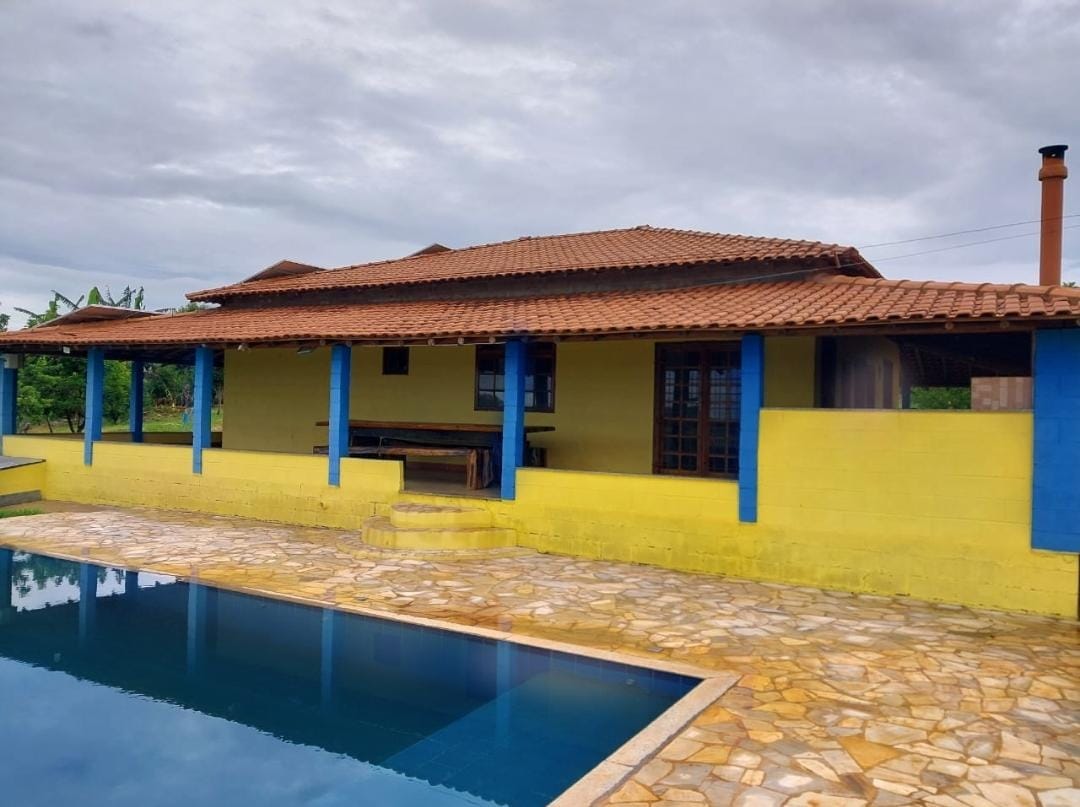 A spacious accommodation features a vibrant yellow exterior with blue accents and a tiled roof. A well-maintained stone patio surrounds a rectangular swimming pool, reflecting the cloudy sky. Lush greenery is visible in the background, enhancing the tranquil outdoor environment.