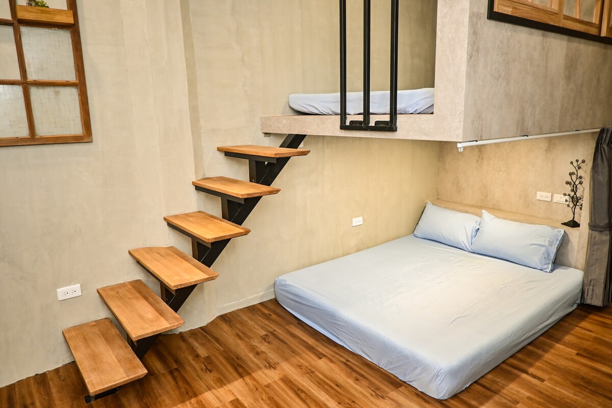 A unique multi-level sleeping area is featured, with a spacious bed positioned on the lower level. A sleek wooden staircase leads to an upper sleeping space with an additional mattress. Neutral-colored walls and warm wood flooring enhance the overall design.