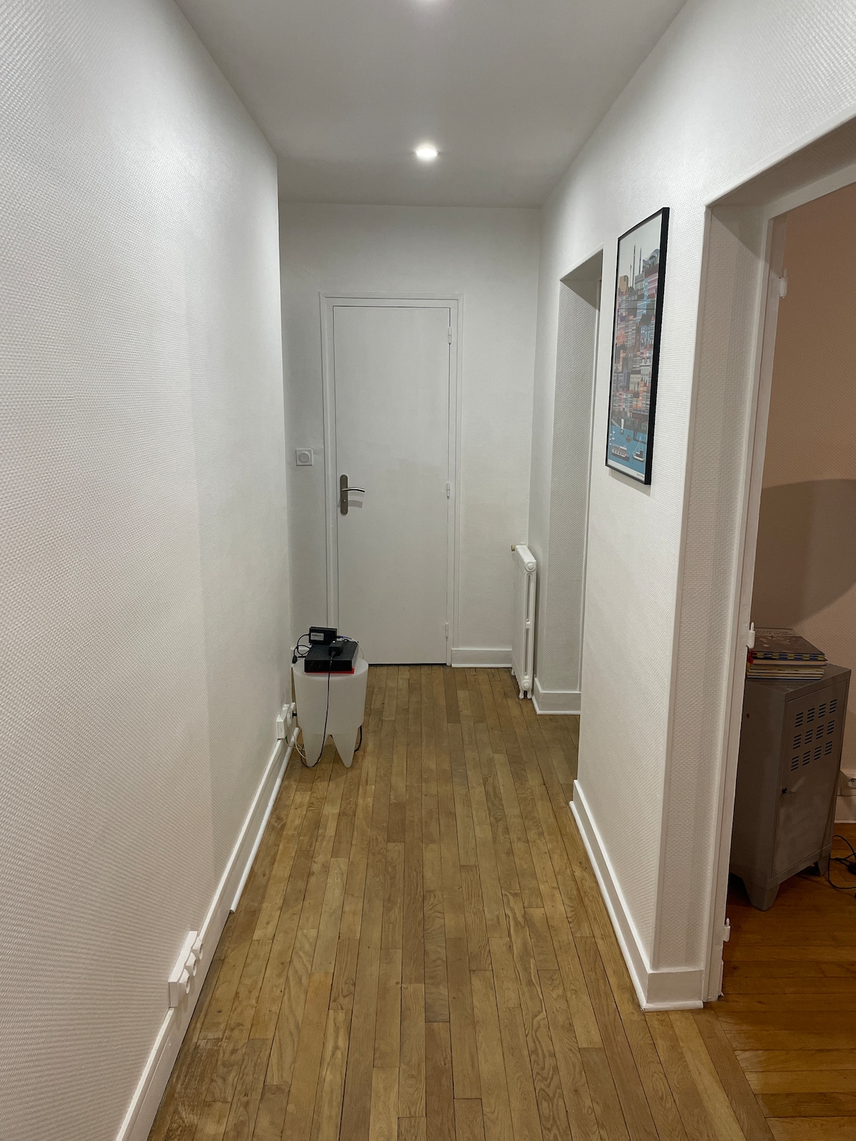 A narrow hallway is illuminated by recessed ceiling lights, showcasing smooth white walls and warm wooden flooring. A closed door leads to another room, and a small side table is positioned beside the wall, enhancing the functional layout of the space.