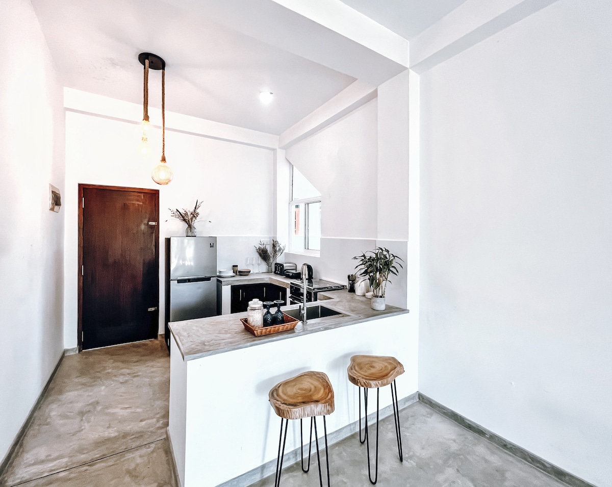 A modern kitchen area features a light countertop with seating for two. Basic cooking appliances and a refrigerator are positioned against the wall. Natural light streams in through a nearby window, illuminating the minimalist decor in shades of white and gray.