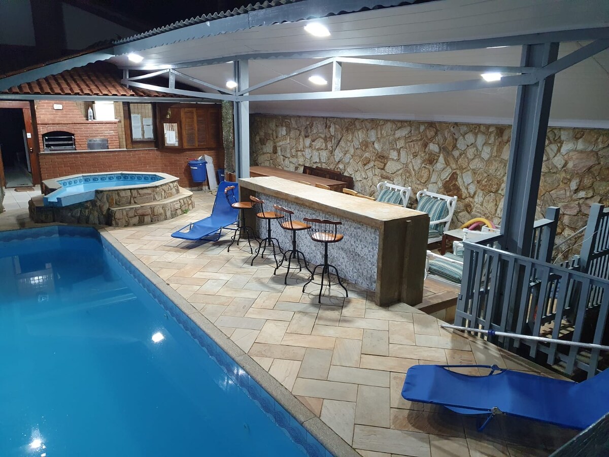A pool area features a clear blue swimming pool beside a warm spa. A stone bar with high chairs is positioned nearby, with lounge chairs situated on the tiled deck. Soft lighting enhances the stone wall backdrop, creating a welcoming space for relaxation.