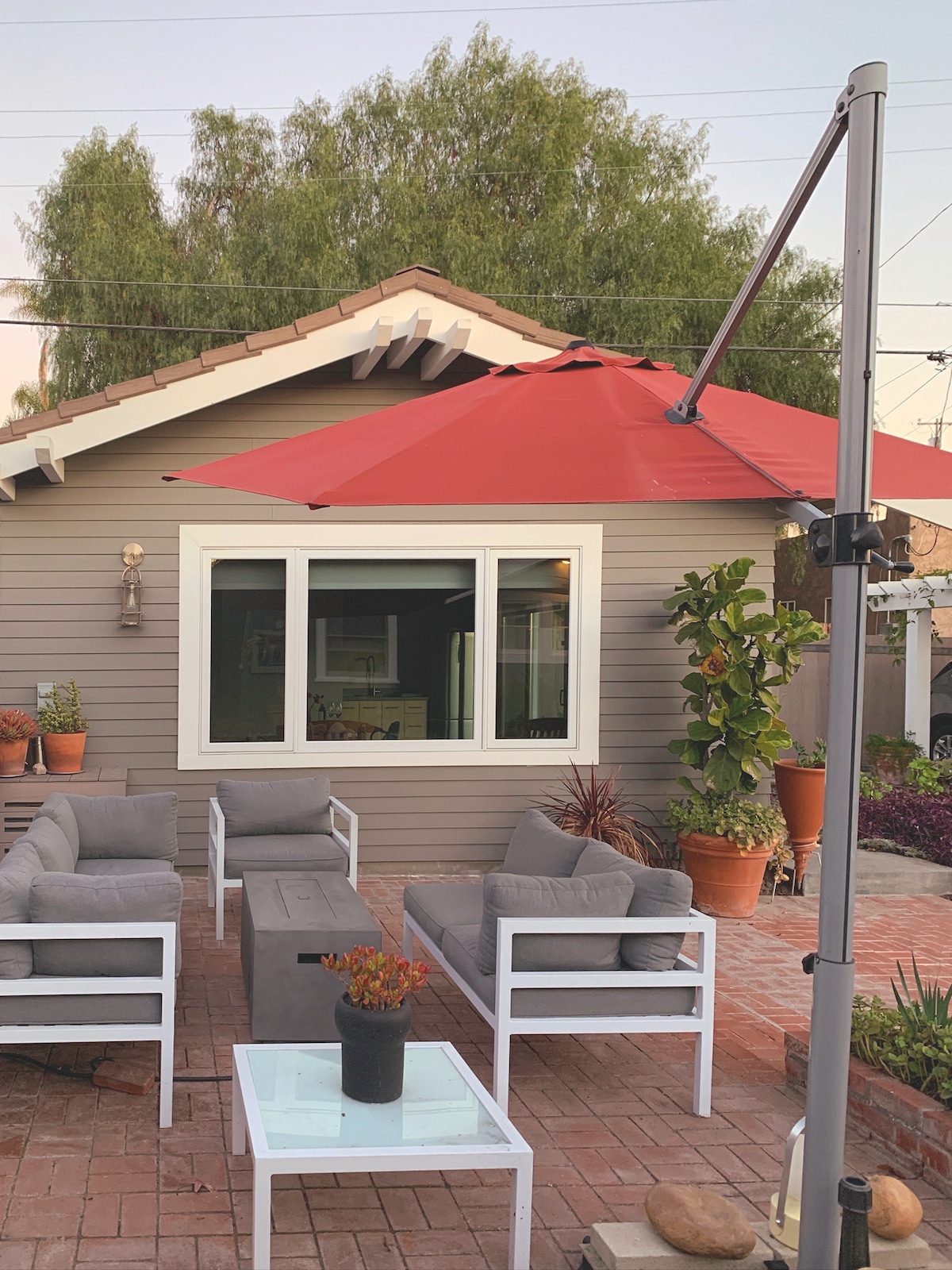 An outdoor patio area is furnished with four gray lounge chairs and a rectangular table in the center. A red umbrella provides shade over the seating arrangement. The backdrop includes a well-maintained structure with a large window and various potted plants. Lush greenery surrounds the setting.