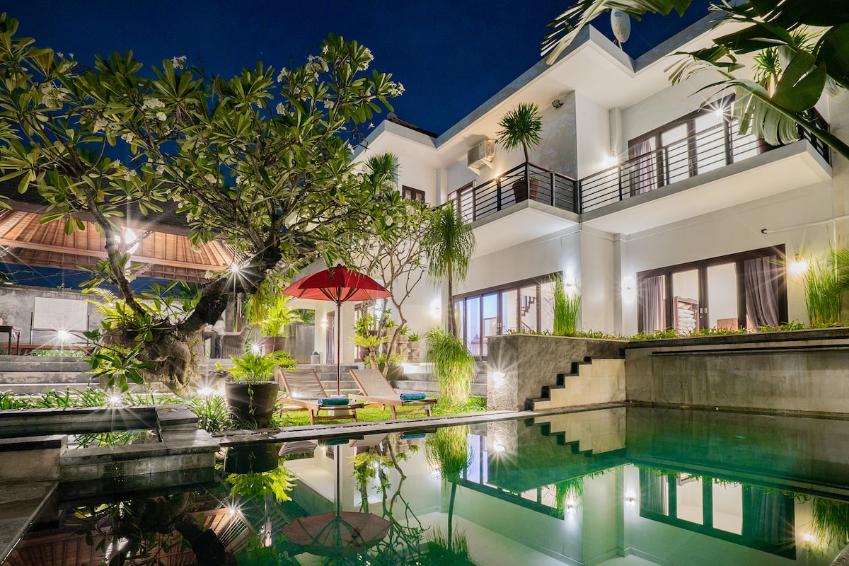 A modern villa is illuminated at night, featuring a tranquil pool reflecting nearby lights. A large tree stands prominently, with vibrant green foliage surrounding the outdoor area. Lounge chairs are positioned near the pool, complemented by a red umbrella, adding a touch of color to the serene setting.