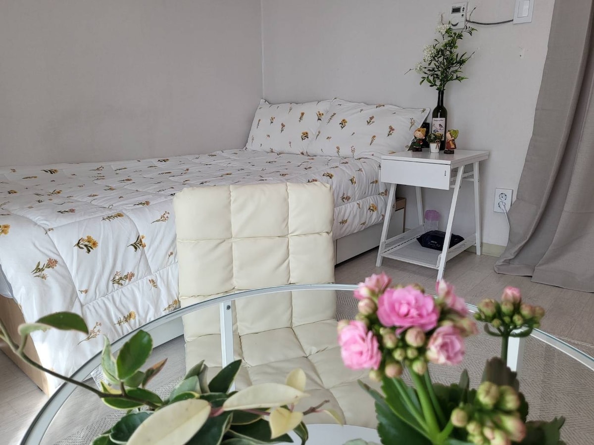 A cozy sleeping area features a comfortable bed dressed in floral-patterned bedding. A small bedside table holds decorative items, while a glass dining table is adorned with fresh flowers, creating a relaxing atmosphere. Soft natural light fills the space through nearby windows.