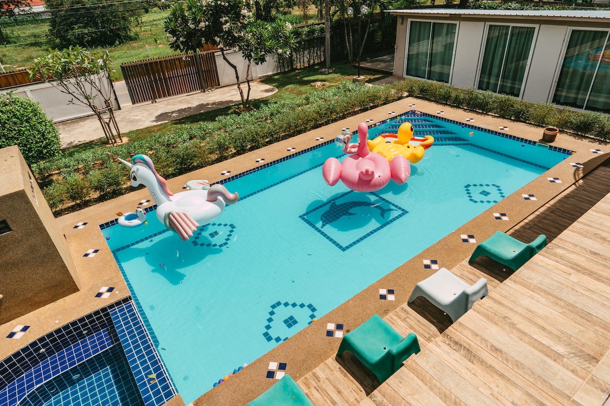 A clear swimming pool features colorful inflatable floats, including a unicorn and a flamingo. Surrounding the pool are neatly arranged loungers and landscaped greenery, offering a serene outdoor space. Views of the villa's exterior can be seen in the background.