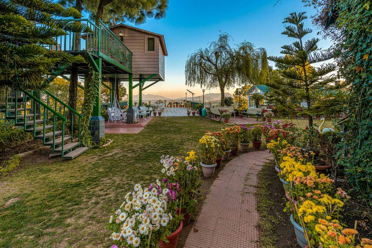 A vibrant pathway lined with potted flowers leads to a patio area featuring a gazebo. The treehouse stands elevated nearby, surrounded by lush greenery. Scenic views are visible in the distance, complemented by the serene setting of the lawn space.