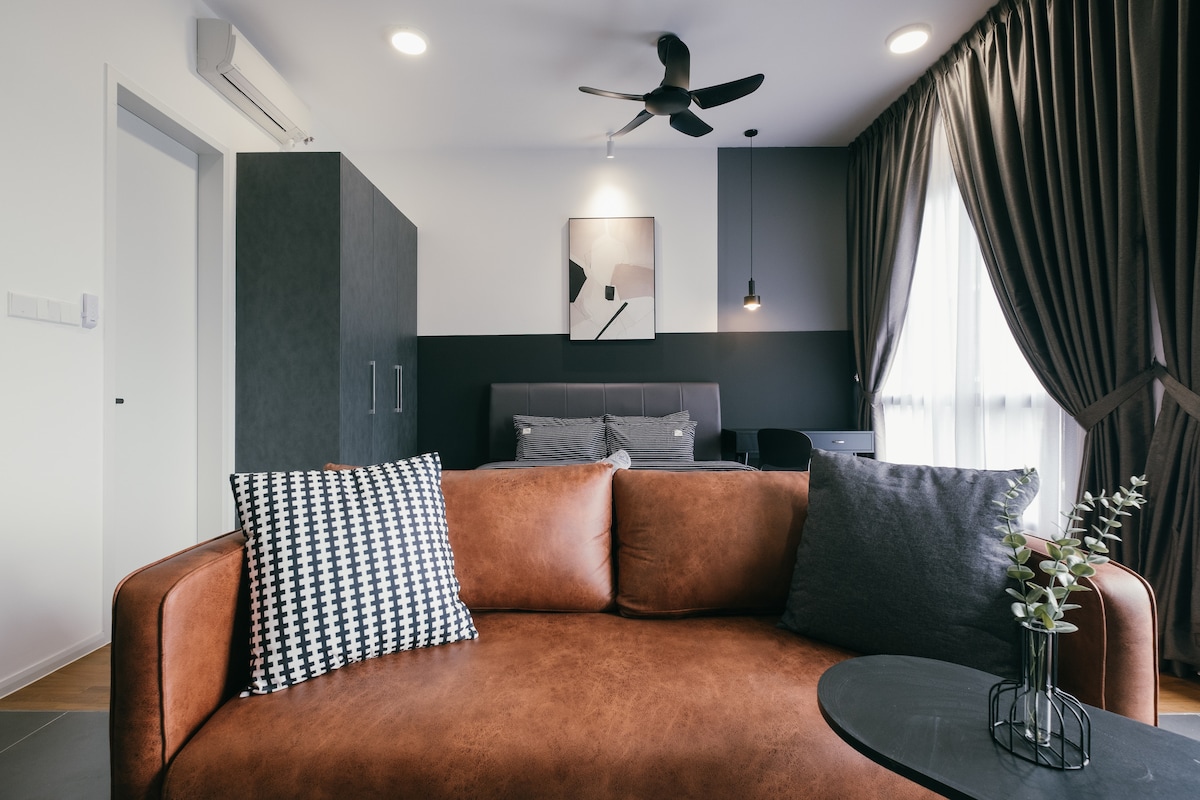 The living area features a brown sofa adorned with patterned cushions, positioned opposite a minimalist black side table. Behind it, a cozy sleeping area is visible, marked by grey bedding and curtains, while a ceiling fan provides ventilation overhead.