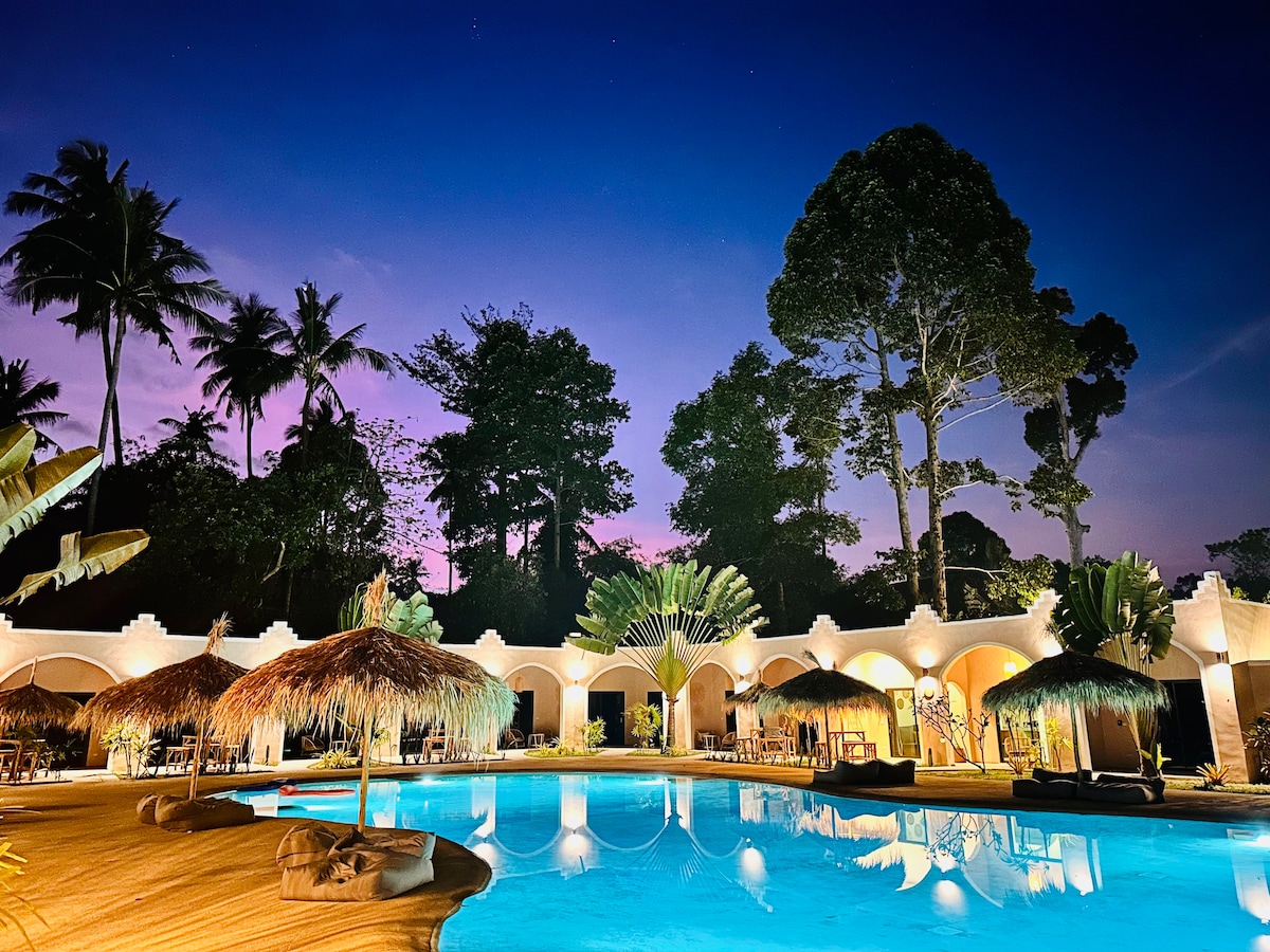 A tranquil pool area is surrounded by lush tropical trees, featuring thatched roof loungers alongside the water. Soft lights illuminate the space, reflecting off the calm surface of the pool. The sky transitions from dusk to night, enhancing the serene atmosphere.