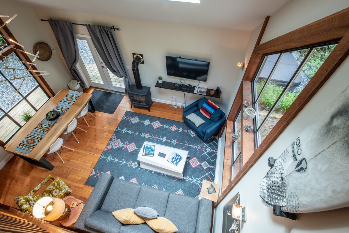 An open living area is seen from above, featuring a cozy sofa and a modern armchair. A dining table sits nearby, surrounded by white chairs. Large windows allow natural light to fill the space, highlighting the hardwood floors and decorative area rug.