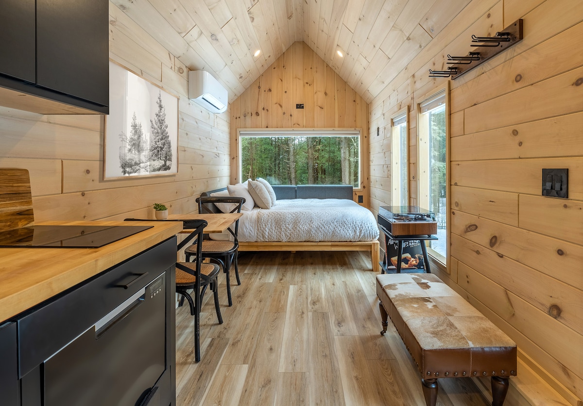 A modern interior is showcased with wooden walls and flooring, featuring a cozy bed positioned near large windows. A compact dining area is visible, with a small table and two chairs. An air conditioning unit is installed on the wall, contributing to the space's comfort.