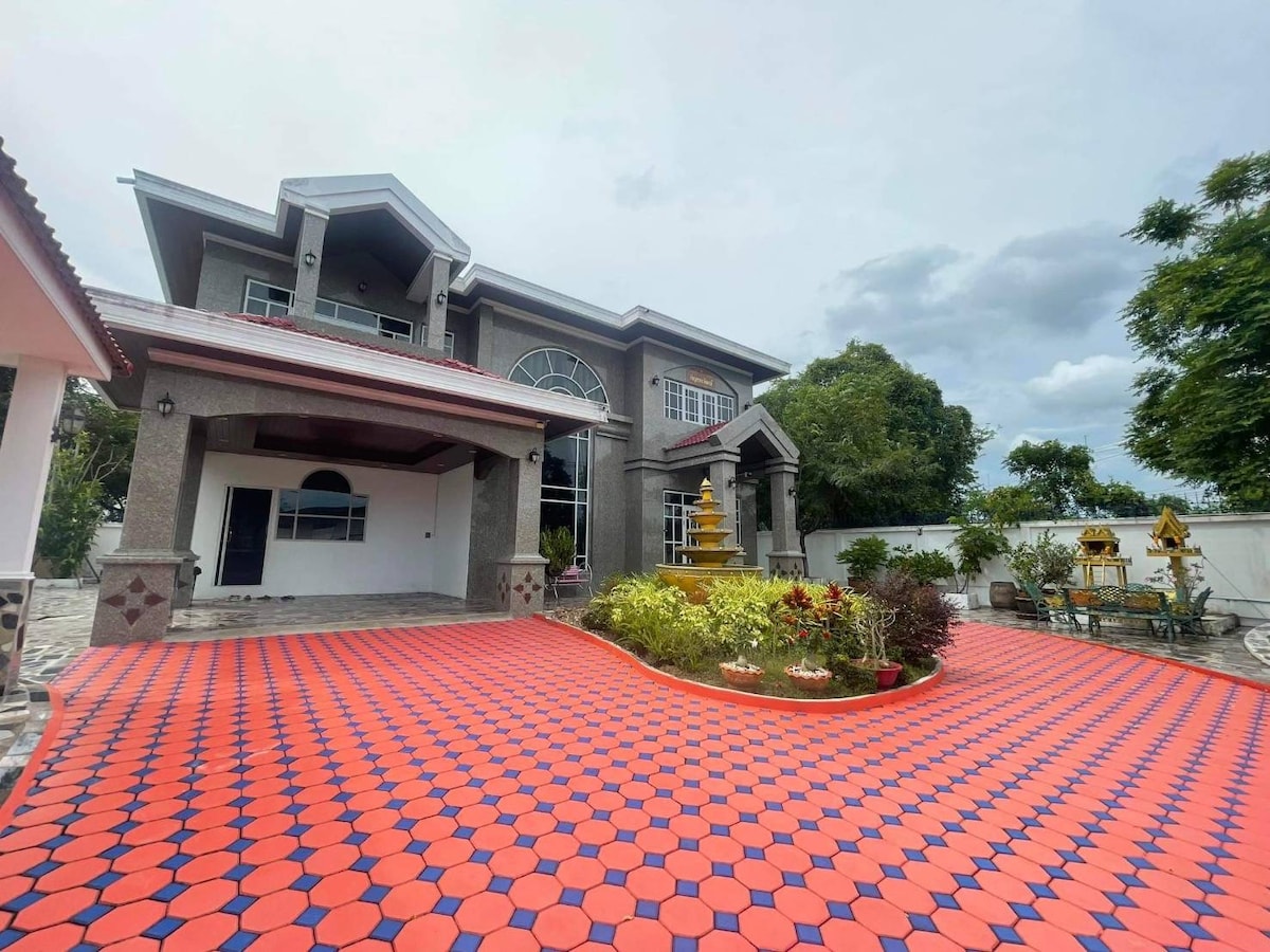 A large, two-story house is framed by a spacious outdoor area featuring a brightly patterned red and blue tiled surface. Lush greenery is arranged in garden beds along the perimeter, while the architecture displays a blend of modern and traditional design elements.