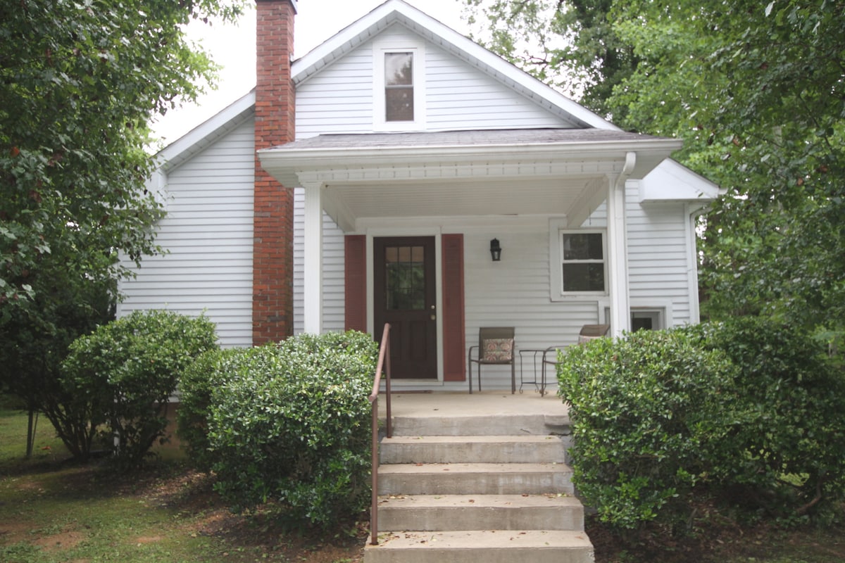 The cottage's exterior showcases a welcoming porch with two chairs positioned on a concrete pathway. Lush greenery surrounds the entrance, and a brick chimney complements the white siding of the home, enhancing its traditional charm.