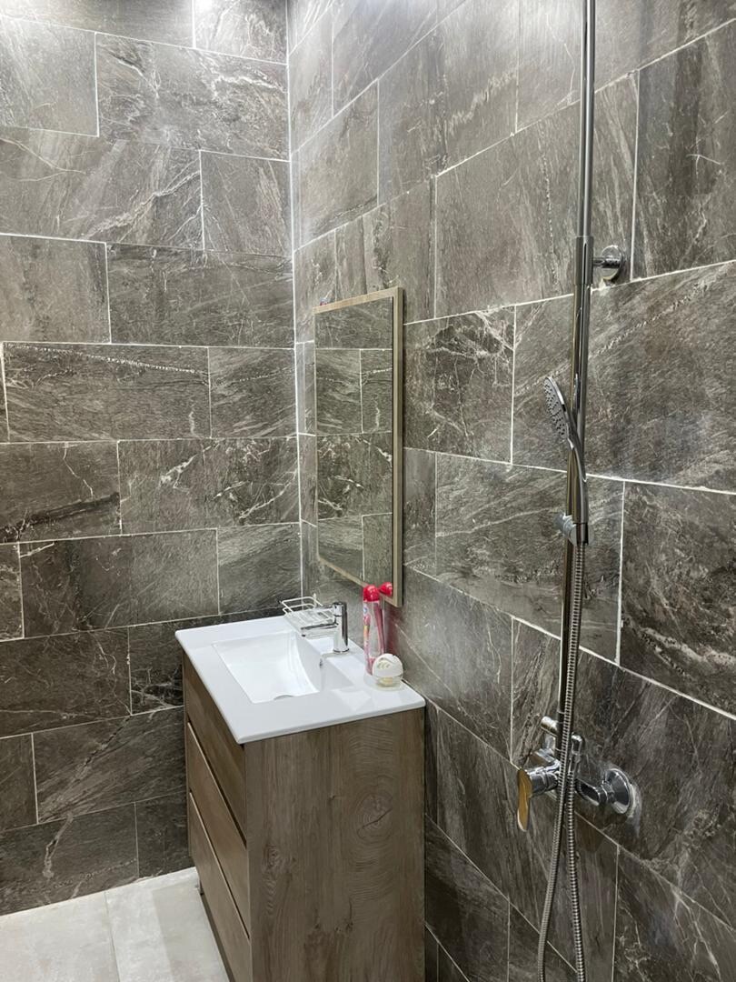 A modern bathroom is displayed, featuring sleek stone tiles in varying shades of gray. A wooden vanity with a white sink is located against the wall, accompanied by a wall-mounted mirror. A glass shower is positioned adjacent, showcasing a stylish showerhead.