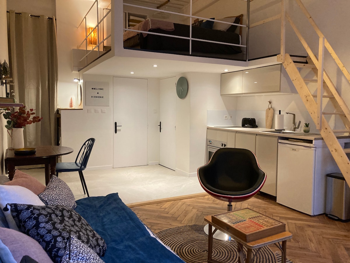 A modern duplex features an open living area with a spacious sofa and a decorative coffee table. The kitchenette is equipped with essential appliances. Above, a cozy mezzanine bedroom is visible, along with a stylish staircase. Natural light fills the space, enhancing the neutral decor.