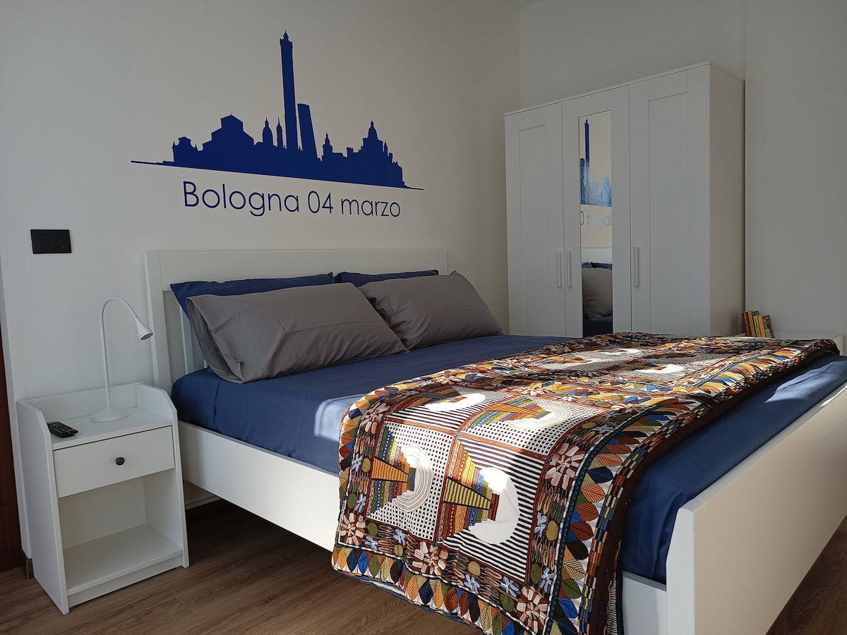 A bright bedroom features a spacious bed adorned with a colorful blanket. A contemporary white nightstand with a lamp stands beside the bed. The wall is decorated with a blue silhouette of Bologna's skyline, complemented by a mirrored wardrobe providing additional storage.