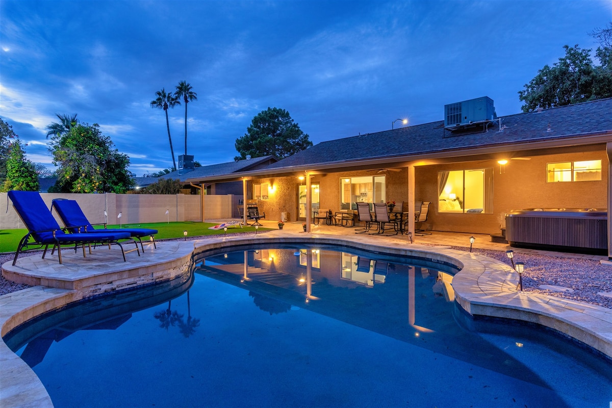 The backyard features a heated pool surrounded by a travertine patio. Lounge chairs are positioned for relaxation, while the exterior of the home is softly illuminated by outdoor lighting at dusk. The setting includes lush greenery and an inviting hot tub.