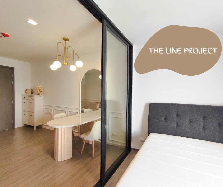 A modern living space showcases a spacious bed complemented by a stylish upholstered headboard. Adjacent to the bed, a dining area features a round table surrounded by contemporary chairs, illuminated by a distinctive light fixture. A glass partition adds an element of separation while enhancing light flow.