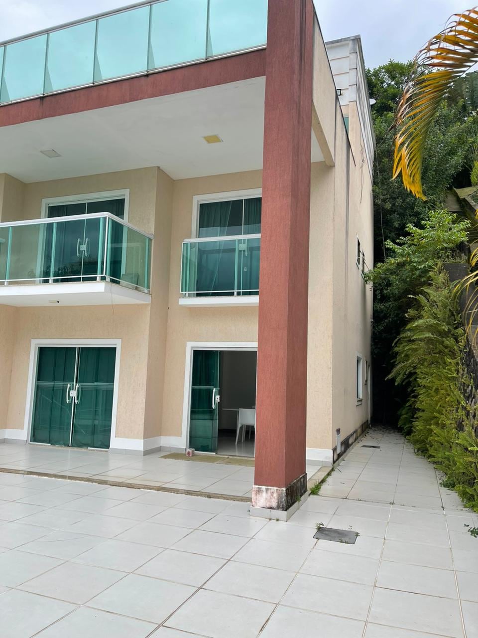The exterior of the building features two levels with glass balconies and large glass doors. A tiled patio surrounds the base, leading to a narrow pathway bordered by greenery. The overall design emphasizes an open and spacious feel.