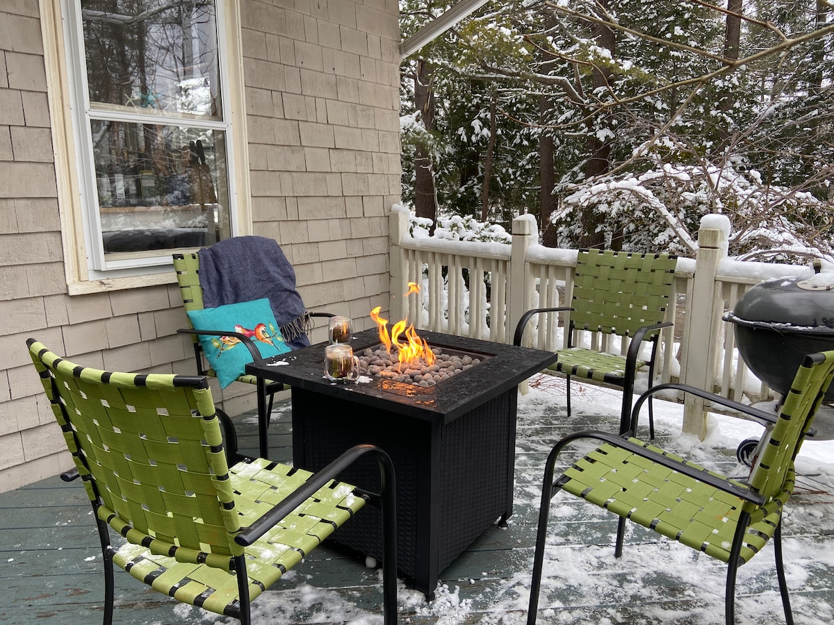 A cozy outdoor space is shown with four green woven chairs arranged around a modern fire pit, which emits a warm flame. Snow blankets the ground, and a blanket rests on one of the chairs, while a grill is positioned nearby.