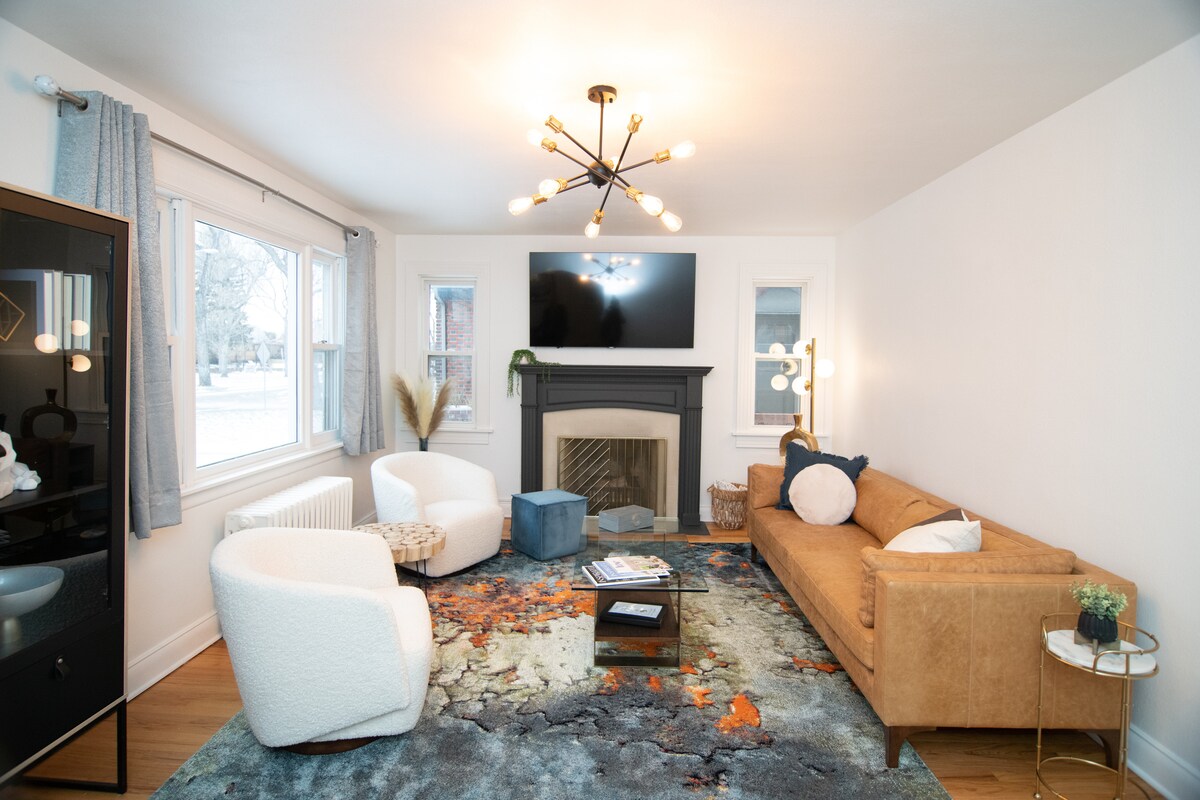 The living area features a modern design with a cozy sofa and two accent chairs positioned around a coffee table. A decorative area rug lies on the hardwood floor, and a large TV is mounted above a stylish fireplace. Natural light filters through multiple windows.