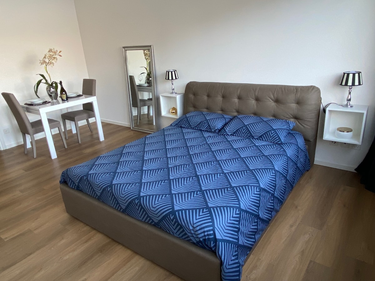A spacious bed covered with a blue patterned comforter is situated in the room. A mirrored dresser reflects the soft lighting. In the background, a dining area with a table and two chairs is visible, accompanied by elegant table decorations.