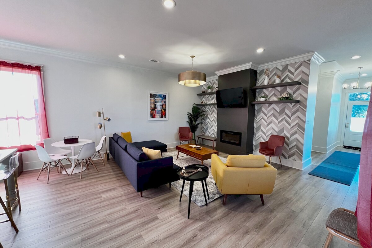 A spacious living area features a mix of seating options, including a yellow sofa and a blue couch, around a central coffee table. A wall-mounted fireplace adds warmth. Bright windows allow natural light, complemented by modern decor and neutral tones.