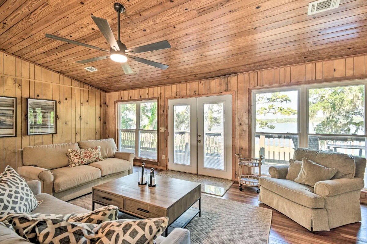 A spacious living area features wooden paneling and a vaulted ceiling, with multiple windows allowing natural light. Comfortable seating is arranged around a central coffee table, while a ceiling fan provides airflow. Double glass doors lead to an outdoor balcony, enhancing the open atmosphere.
