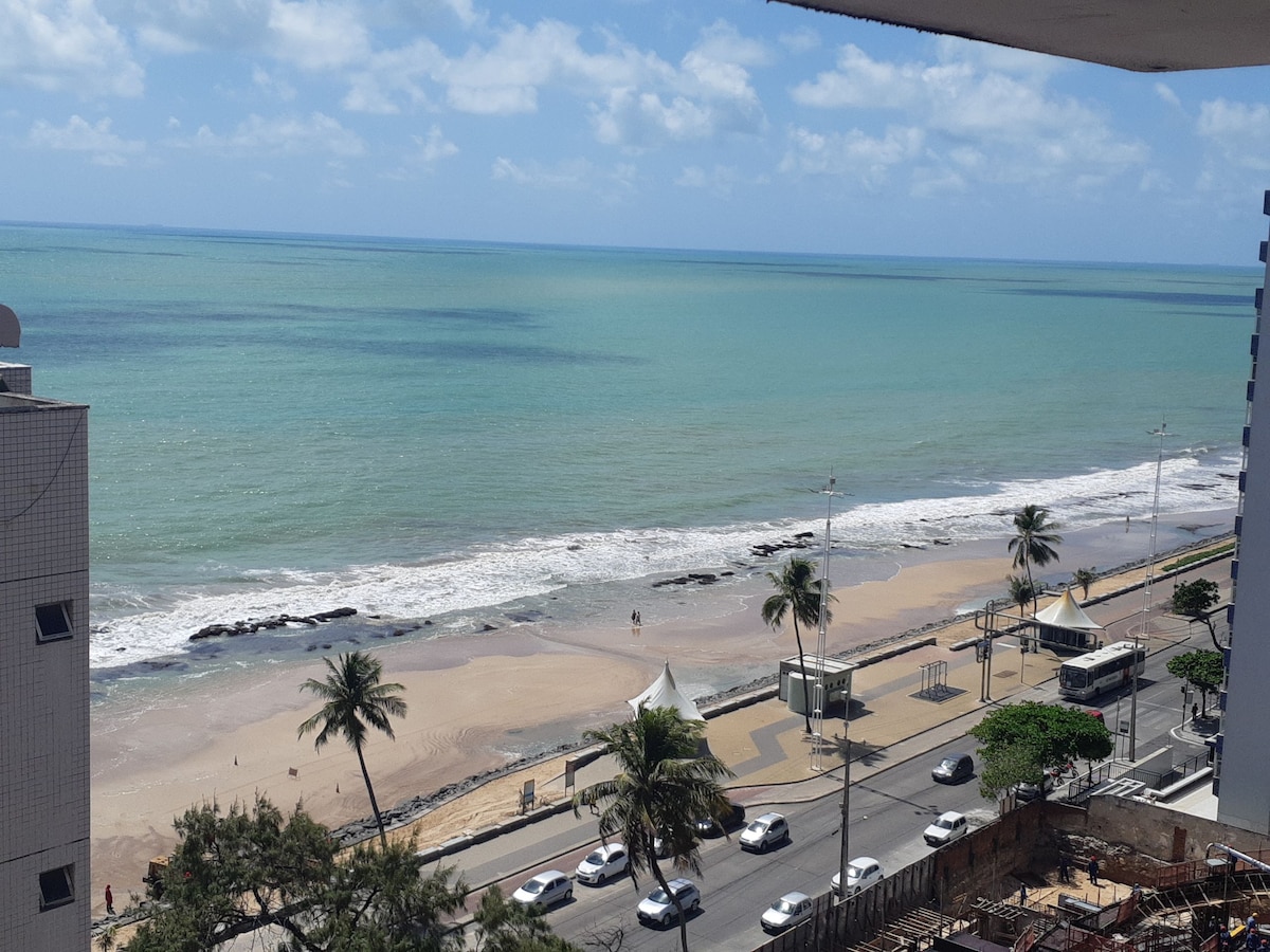 A panoramic view of the sea is visible, showcasing varying shades of blue and green water. The sandy beach is partially adorned with gentle waves. Palm trees line the shore, and a coastal road with vehicles is seen in the foreground.