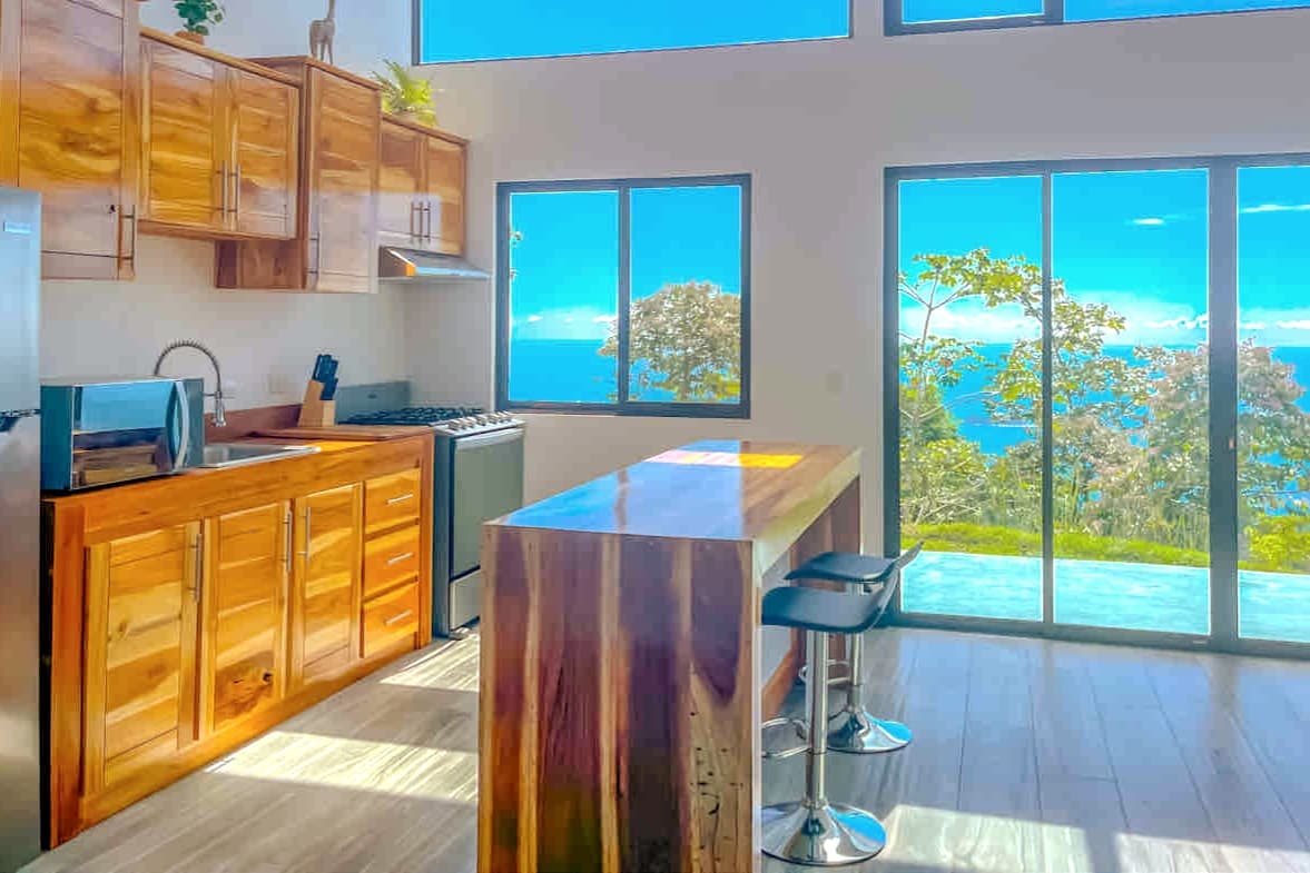 A modern kitchen is displayed, featuring wooden cabinetry and an island counter with stools. Large windows offer a panoramic view of the ocean and surrounding jungle, allowing natural light to fill the space. Stainless steel appliances are positioned for convenience.