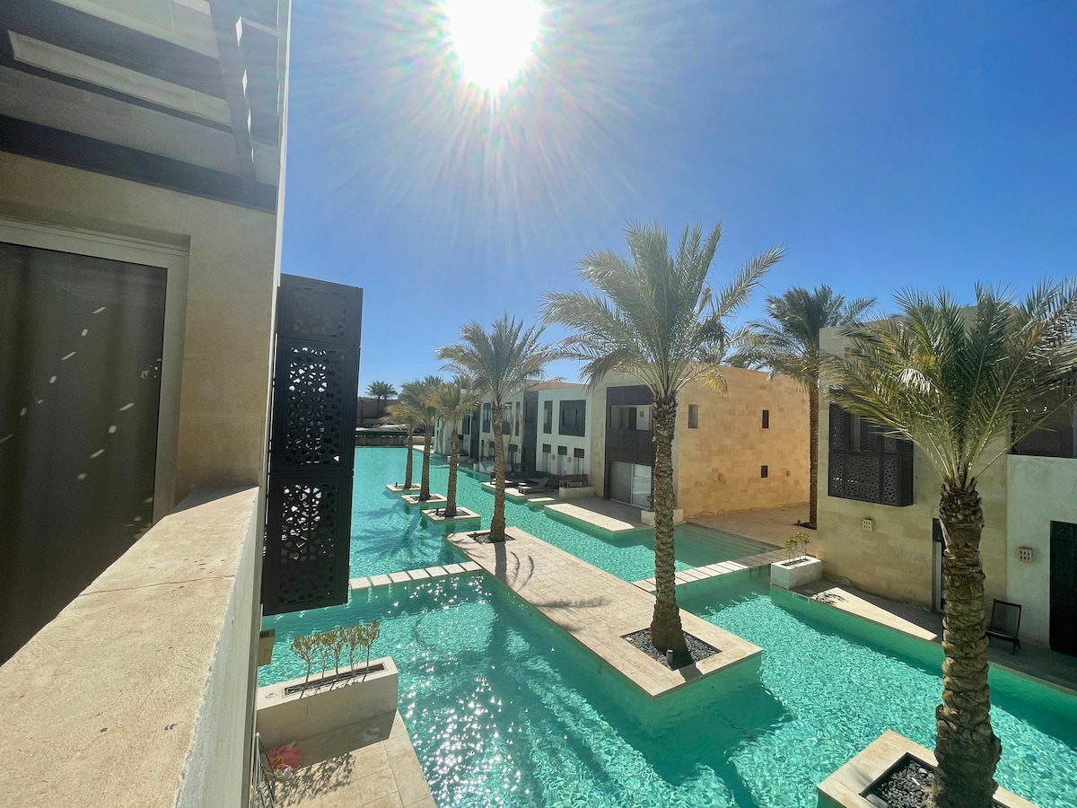 A bright view of the pool area is captured, featuring palm trees lining the water’s edge. The sun shines brightly overhead, casting reflections on the turquoise water. Surrounding buildings showcase a harmonious blend of stone and modern architecture.