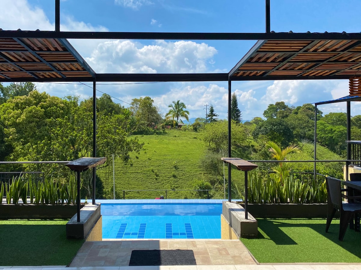 An outdoor pool area is visible, framed by modern architectural elements. Lush greenery surrounds the space, with distant hills and trees seen in the background. Glass railings and stone accents contribute to a contemporary feel, creating an inviting atmosphere for relaxation.