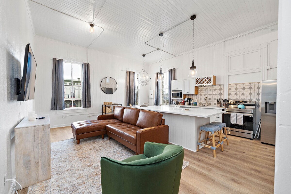 A spacious open living area is presented, featuring a comfortable brown leather sectional sofa and a green accent chair. Large windows with sheer curtains allow natural light to brighten the room. The kitchen includes modern appliances, a breakfast bar, and decorative tile backsplash.