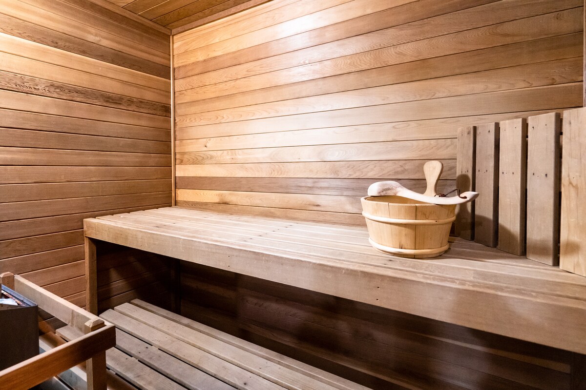 The sauna is constructed with smooth wooden panels, providing a warm environment. A wooden bucket and ladle are placed on a bench, which offers seating along two sides. Adjacent benches add additional space for relaxation.